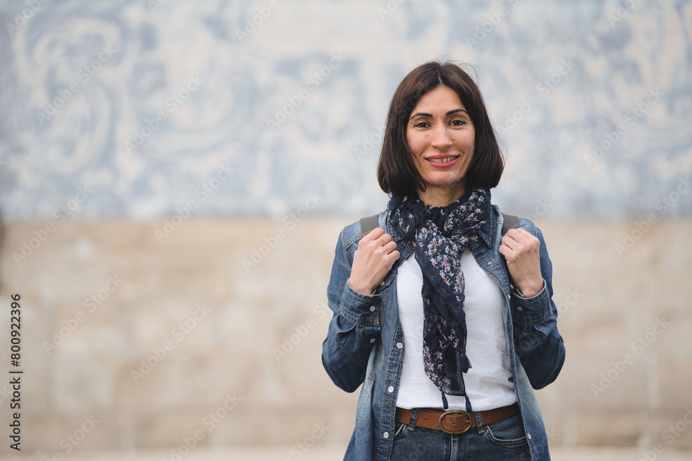 Portrait of female tourist standing with scarf and backpack smiling looking at camera near church with famous Portuguese blue ceramic tiles. Copy space.