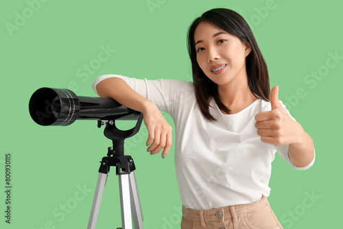 Young Asian woman with telescope showing thumb-up on green background