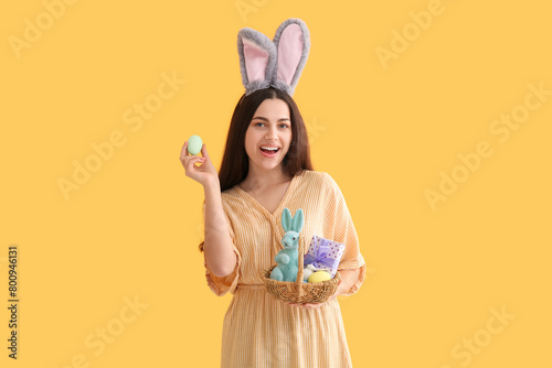 Beautiful young happy woman in bunny ears holding basket with Easter painted eggs and gift box on yellow background