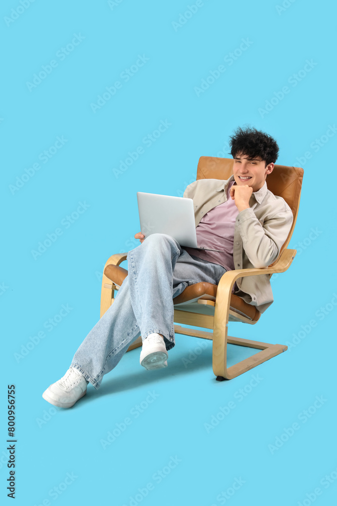 Young man resting on comfortable armchair with laptop on blue background