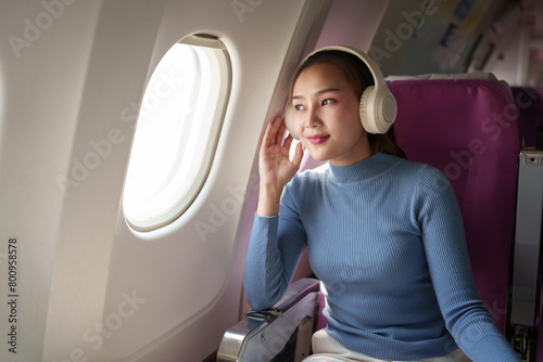 Young woman wearing headphones listening to music while traveling Sitting near the window in First Class during the flight. Traveling. Connect to the WiFi Internet with your smartphone on the plane.