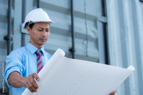 An engineer wearing a hard hat and holding a blueprint is looking at the construction site.
