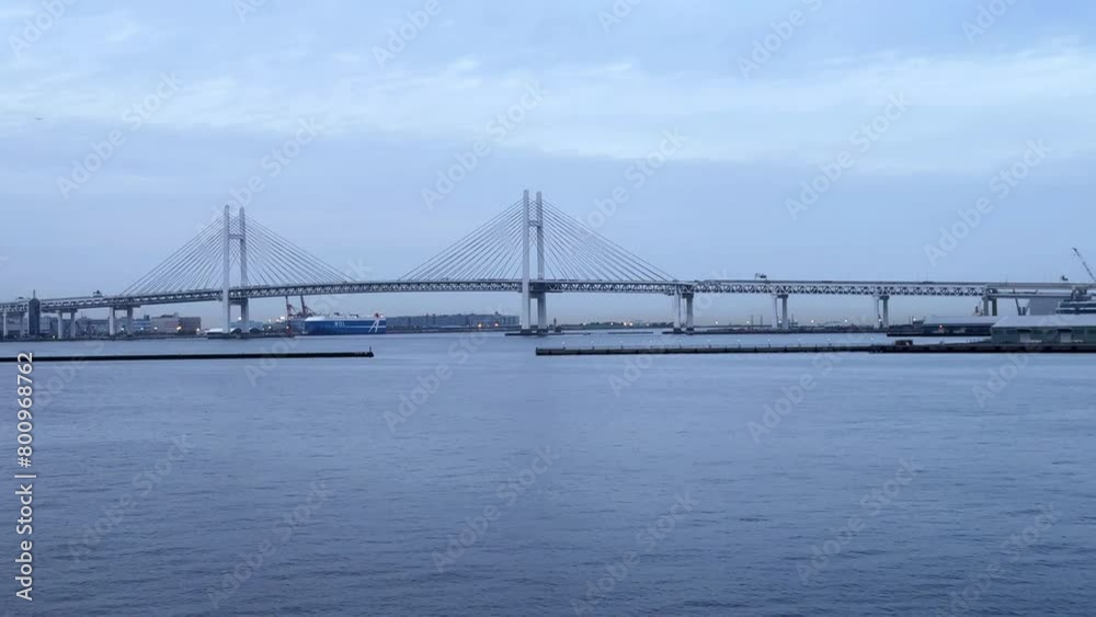 Distant view of a suspension bridge over serene water with boats, under a cloudy sky