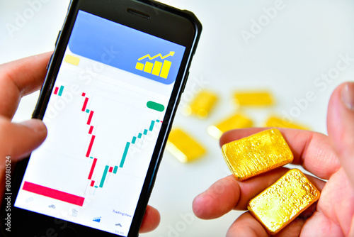 Hand holding a smartphone and a gold bar to check the gold price graph for investment