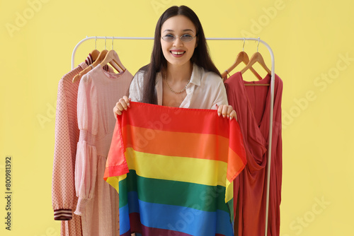 Female Asian fashion designer with LGBT flag and clothes rack on yellow backg...