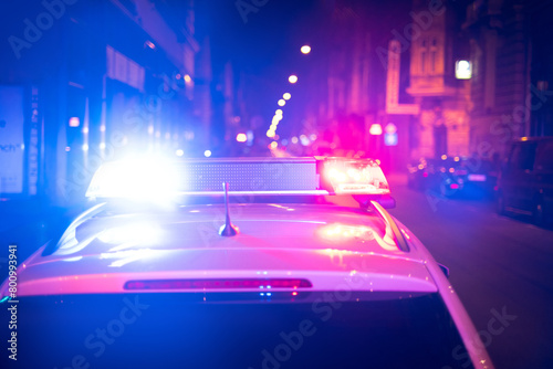 A POLICE CAR IS STANDING AT THE SCENE OF THE INCIDENT IN THE CITY WITH FLASHING AND SIRENS ON. Blurred old houses in the background.