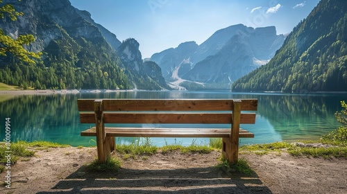 There is a wooden bench in the foreground of the picture. Behind the bench is a lake and in the background are snow-capped mountains. The sky is blue and there are some clouds in the sky.