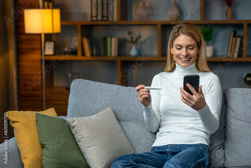 Smiling and excited adult woman checking her recent pregnancy test, sitting on gray couch at home