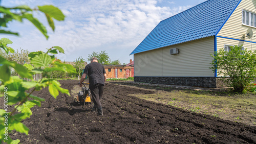 Cottage, plot of land in front of the house. Man ploughing land with cultivator on sunny spring day. Concept of gardening, gardening, dacha photo