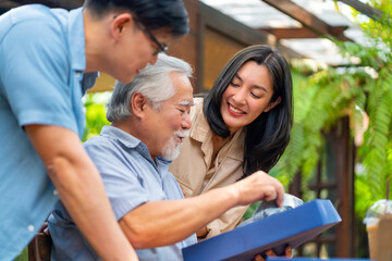 Asian adult couple surprised elderly father with gift box at outdoor cafe restaurant on summer holiday vacation. Family relationship, celebrating father's day and senior people health care concept.