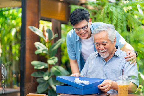 Asian man surprised elderly father with Birthday gift at outdoor cafe restaurant on summer holiday vacation. Family relationship, celebrating father's day and senior people health care concept. photo