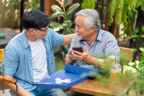 Happy Asian man teaching elderly father using mobile phone application during having lunch together at restaurant on summer vacation. Family relationship senior people mental health care concept.