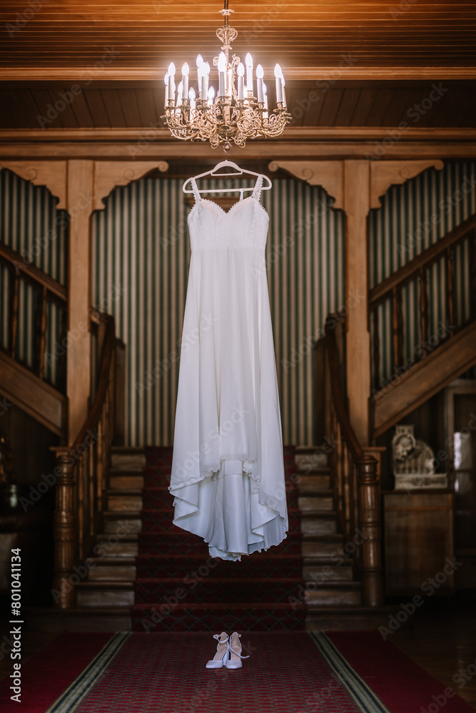 Valmiera, Latvia - August 19, 2023 - A bridal gown suspended from a chandelier in a luxurious wooden hallway with symmetrical staircases, enhancing its elegance. Valmiera, Latvia - August 19, 2023 - A bridal gown suspended from a chandelier in a luxurious wooden hallway with symmetrical staircases, enhancing its elegance.