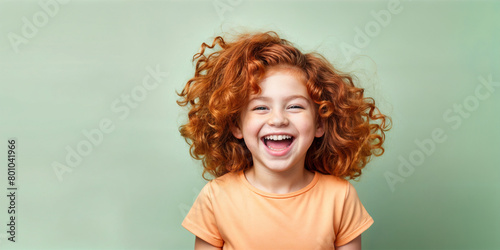 Cute young child  Little girl smiles. Emotion and child development. Little girl smiles while red hair.  A young girl with curly hair is smiling and laughing while wearing a shirt. © Igor