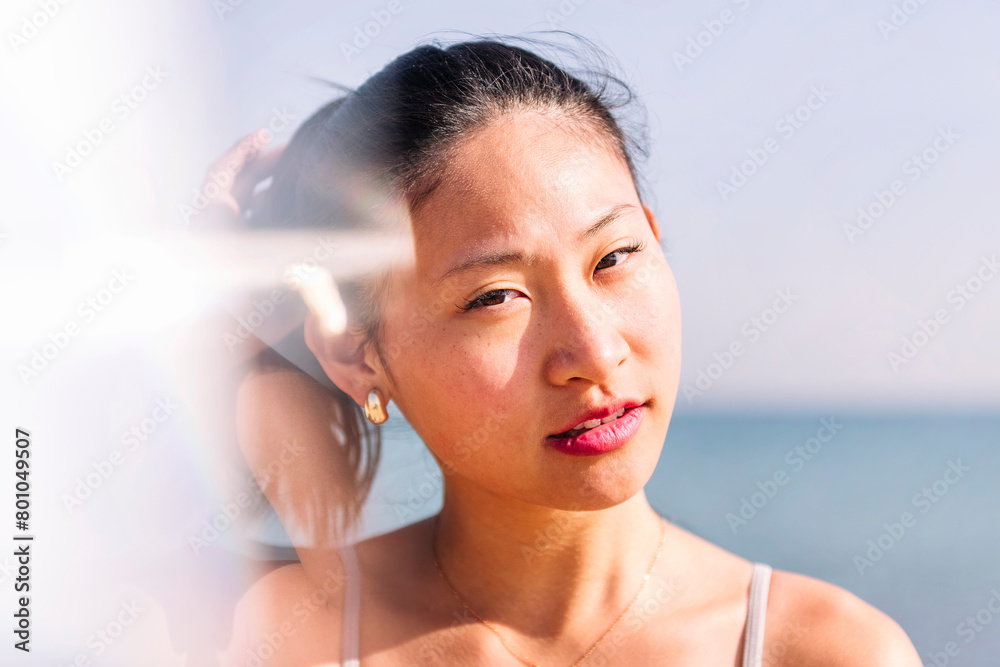 Naklejka premium young contemplative asian woman at beach looking at camera, concept of beauty and purity, flare effect