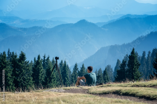 Rear view of a man sitting on the grass while taking pictures with camera on tripod of picturesque mountain slopes of Rhodopi Mountains, Bulgaria.