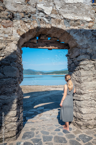 Rear view of young long-haired woman in а dress, leaning against old stone arch enjoying view of beautiful lake. © Jess_Ivanova