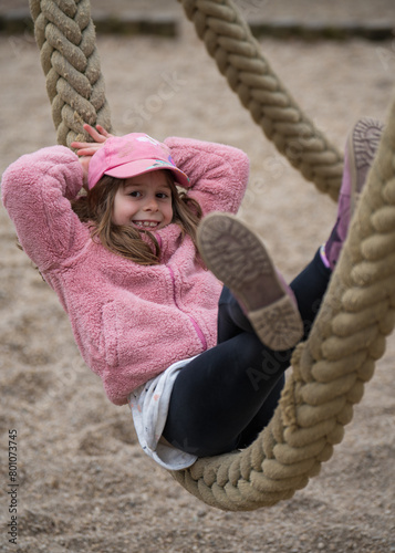 A joyful, happy girl of 7 years old plays happily in nature in a city park. Lying on a thick rope, on a carousel.  child is a cap