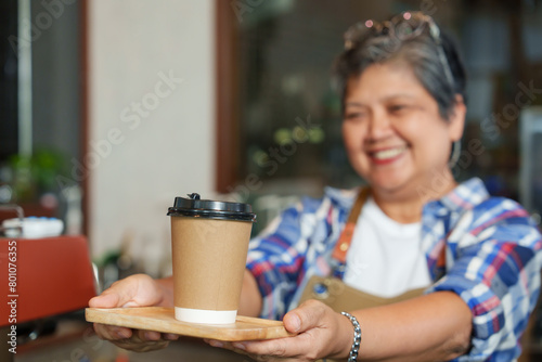 Smiling senior barista offering a takeaway coffee cup  service with personal touch in cozy cafe Cheerful mature barista in a plaid shirt serving a fresh brew in a paper cup with friendly gesture.