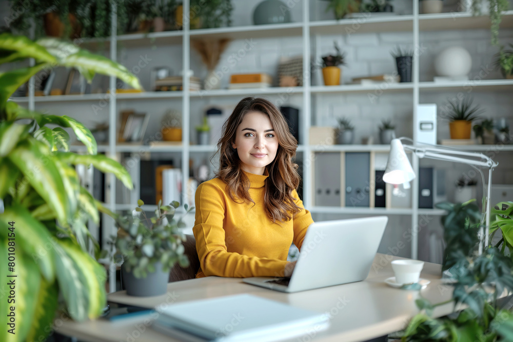 Fototapeta premium Smiling woman, dressed in yellow clothing in an office with laptop, sitting at a desk with white shelves in the background