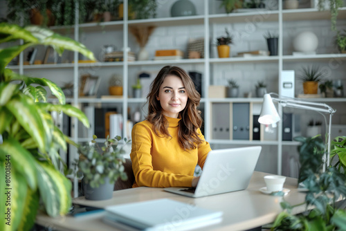 Smiling woman, dressed in yellow clothing in an office with laptop, sitting at a desk with white shelves in the background