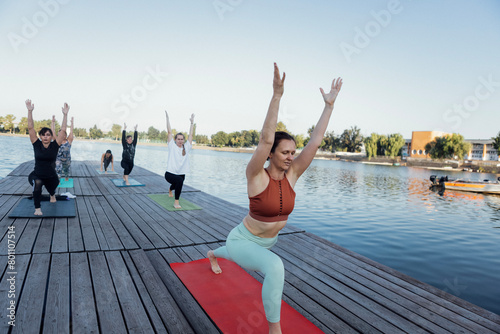 Morning yoga class with an instructor at the pier on the river. Young women stand in a warrior pose on mats and do Virabhadrasana.