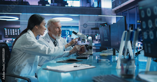 Biotechnology Medical Research Center: Female Asian Neuroscientist Using Desktop Computer And Talking To Male Caucasian Doctor With Microscope. Specialists Developing Innovative Cancer Treatment.