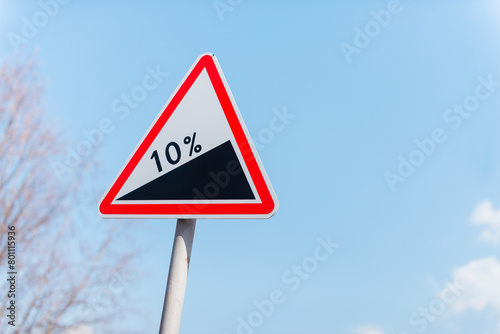 Red triangle road sign indicating a steep 10 percent downhill gradient in the road ahead. Road sign on a background of blue sky