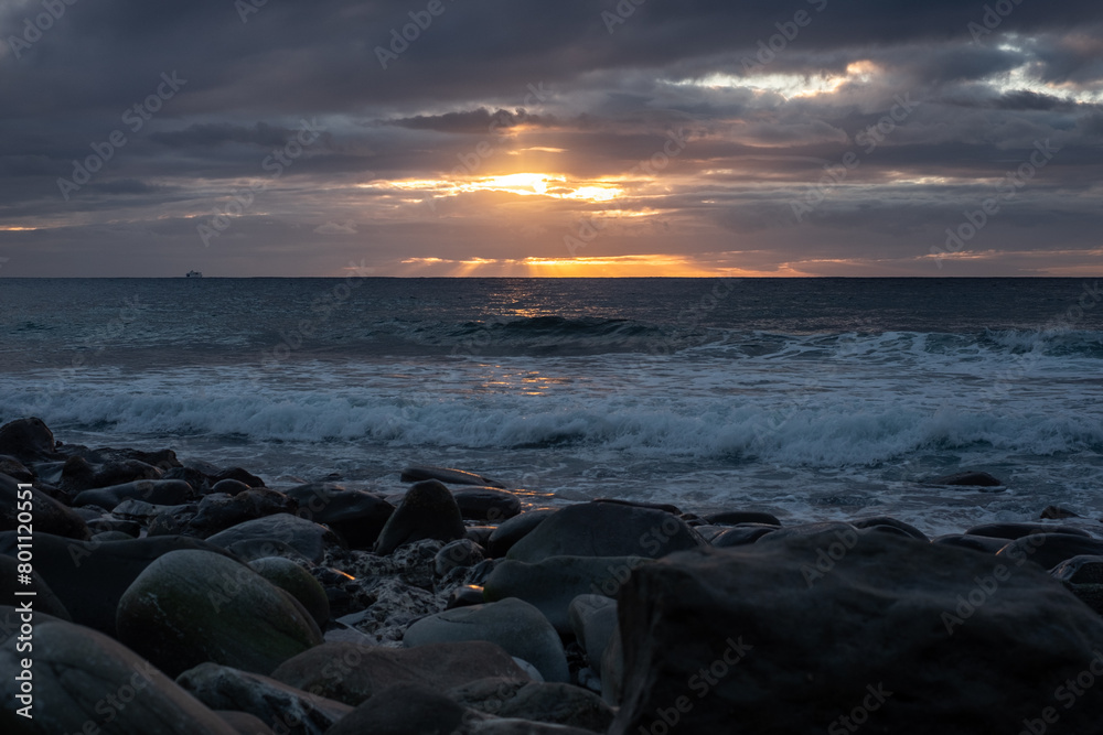 Spectacular view of the cloudy sky at sunset over the Atlantic Ocean, golden rays of sun coming out of the clouds touching the horizon, the pebble beach and the crashing waves, south of Tenerife,
