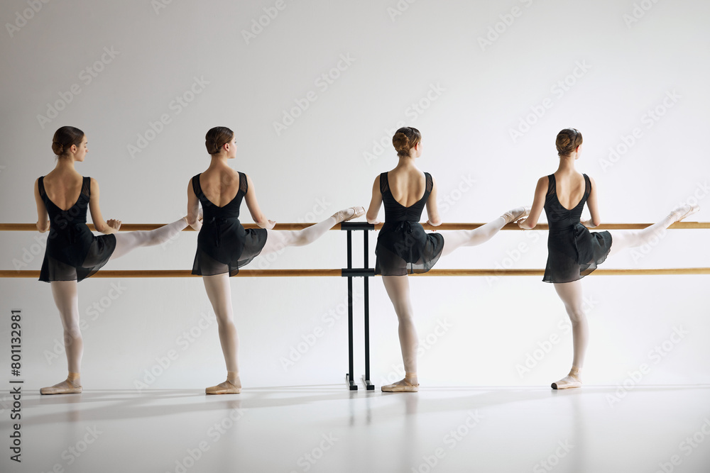 Fototapeta premium Young girls, ballet dancers in black attire and pointe standing next to barre and training, practicing against grey studio background. Concept of ballet, art, dance studio, classical style, youth