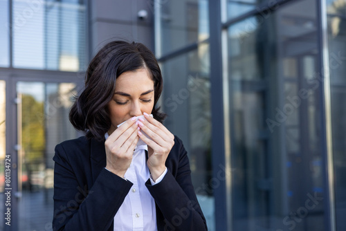 A close-up photo of a sick young woman in a business suit standing outside a building and wiping her nose with a napkin from a runny nose