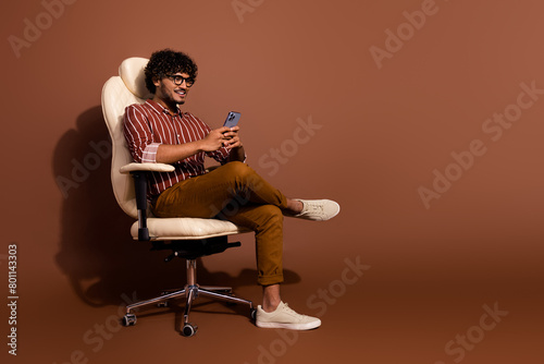 Full size photo of nice young man sit chair smart phone empty space wear shirt isolated on brown color background