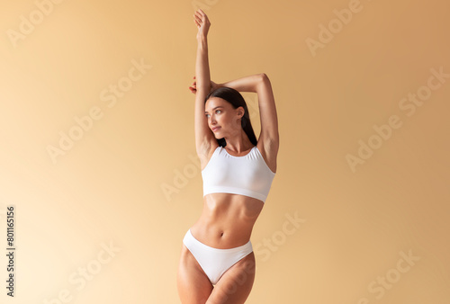 Perfect body. Slim caucasian lady posing with hands raised up, posing in white lingerie at beige background
