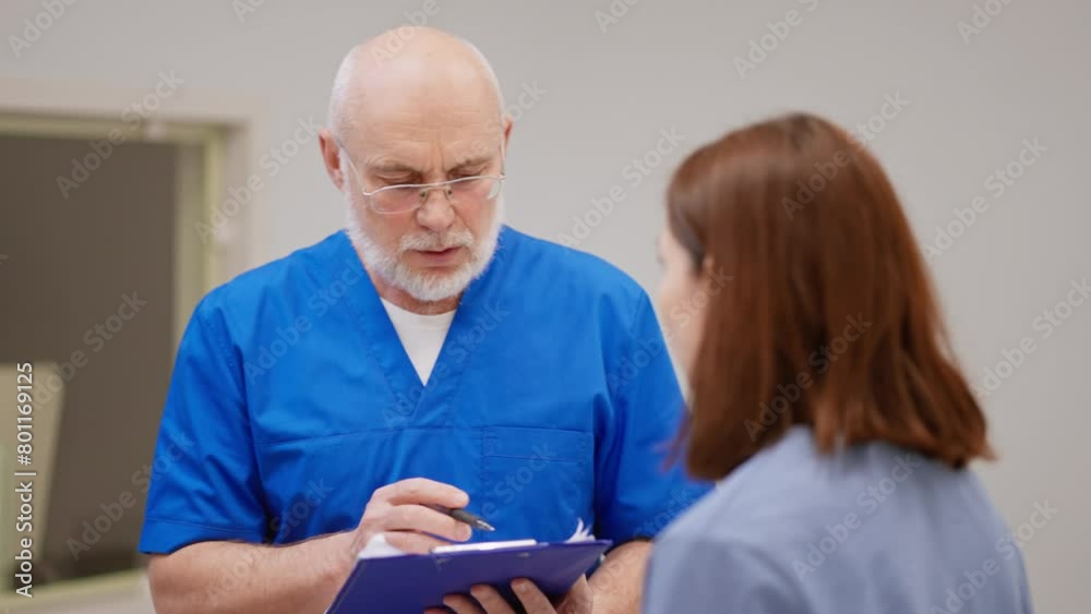 A confident man with glasses an elderly doctor in a blue uniform holds a blue tablet in his hands and talks with a brunette girl about her problems of disease prevention and diagnoses in a modern A confident man with glasses an elderly doctor in a blue uniform holds a blue tablet in his hands and talks with a brunette girl about her problems of disease prevention and diagnoses in a modern