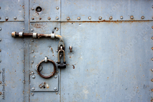 A fragment of an old gate, which is covered with iron. A rusty padlock hangs on an iron bolt. Ring handle on an old door with rivets on iron sheets. The door is locked. A rough lock on the door