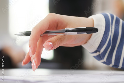 Schoolgirl or student holding a pen in her hand - writing a dictation, preparing an answer at an exam or filling out documents in the classroom, sitting at a desk. Photo. Selective focus