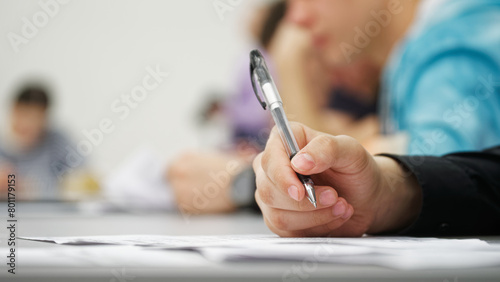 Guy student or schoolboy, writing a dictation, essay or filling out documents in the classroom, sitting at a desk next to other students. Photo. Selective focus. Close-up