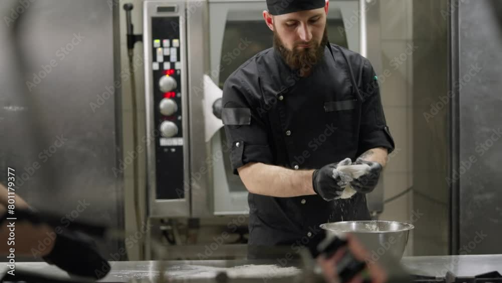 Confident professional chef in a black uniform with a beard places dough on the table with flour and rolls it out with his hands while preparing pizza in the kitchen in a restaurant Confident professional chef in a black uniform with a beard places dough on the table with flour and rolls it out with his hands while preparing pizza in the kitchen in a restaurant