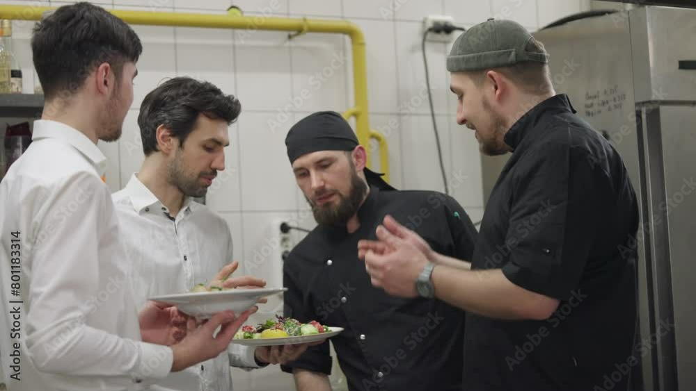 Two chefs in black uniforms together with waiters in white shirts communicate about the prepared dishes and solve their problems while working in the kitchen and restaurant. Cooks and waiters solve Two chefs in black uniforms together with waiters in white shirts communicate about the prepared dishes and solve their problems while working in the kitchen and restaurant. Cooks and waiters solve