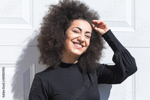 Young white woman with afro-style hair, dressed in black, leaning against a white door, smiling, wearing a headband, facing the sun.