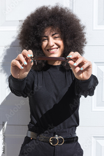 Young white woman with afro-style hair, dressed in black, leaning against a white door, smiling, showing a headband, facing the sun.