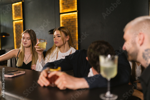 Blonde female friends with cocktails sit near drunk male patrons of bar. Men in black shirts drink strong alcohol laughing