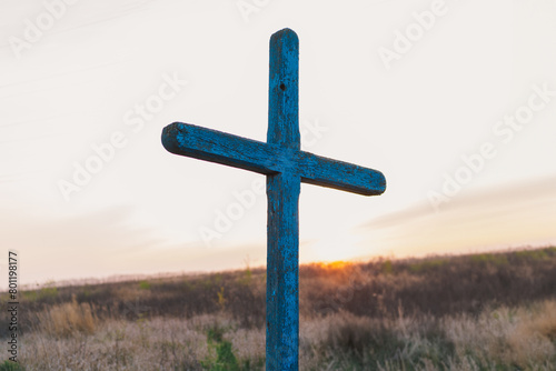 A blue wooden cross is prominently positioned in a field of tall, wild grasses, with the warm glow of the setting sun creating a serene backdrop that evokes a sense of tranquility and reflection.