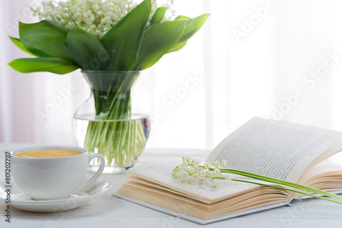 A bouquet of lilies of the valley, cup of morning cappuccino and book on the table. Weekend morning. Selective focus.
