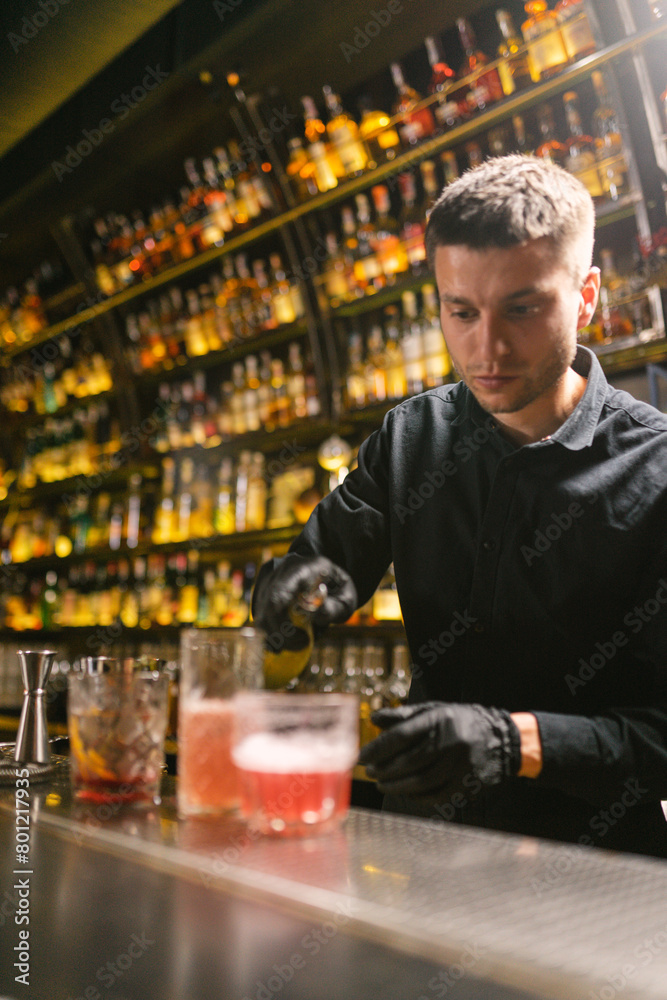 Concentrated barkeeper pours syrup to alcoholic cocktails standing by bar counter. Skilled worker prepares beverages for serving to clients