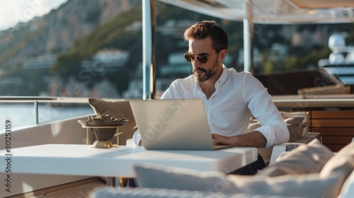 A mid-age male working on laptop computer on deck of a luxury yacht. Remote working concept.