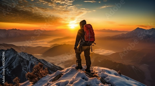 A lone hiker stands on a summit and gazes at the sunrise over a beautiful mountain landscape with clouds and snow