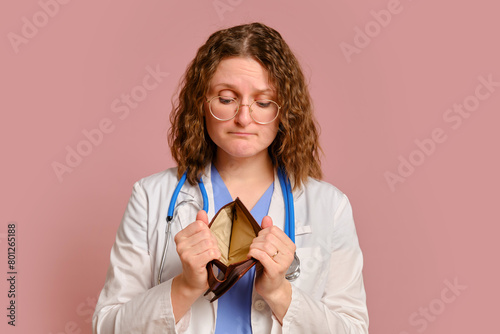 Woman doctor holding an empty wallet in her hands, studio pink background. Nurse in uniform with stethoscope on red studio background © Андрей Журавлев