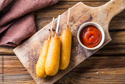 Corn dogs or sausage in roll with ketchup. Top view table scene over a wood background.