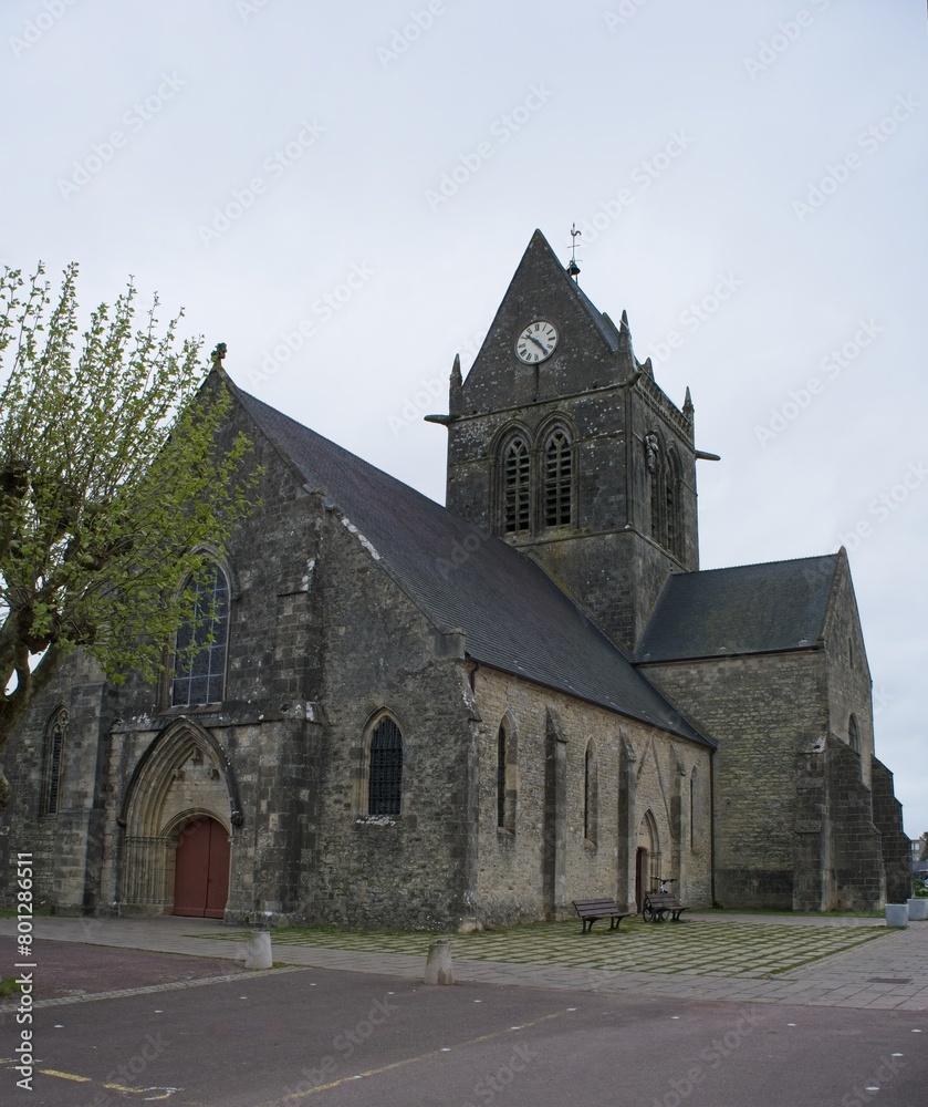 Sainte-Mere-Eglise, France - Apr 19, 2024: Sainte-Mere-Eglise church. People walking in Sainte-Mere-Eglise. Streets and buildings. Lifestyle in the urban area. Cloudy spring day. Selective focus